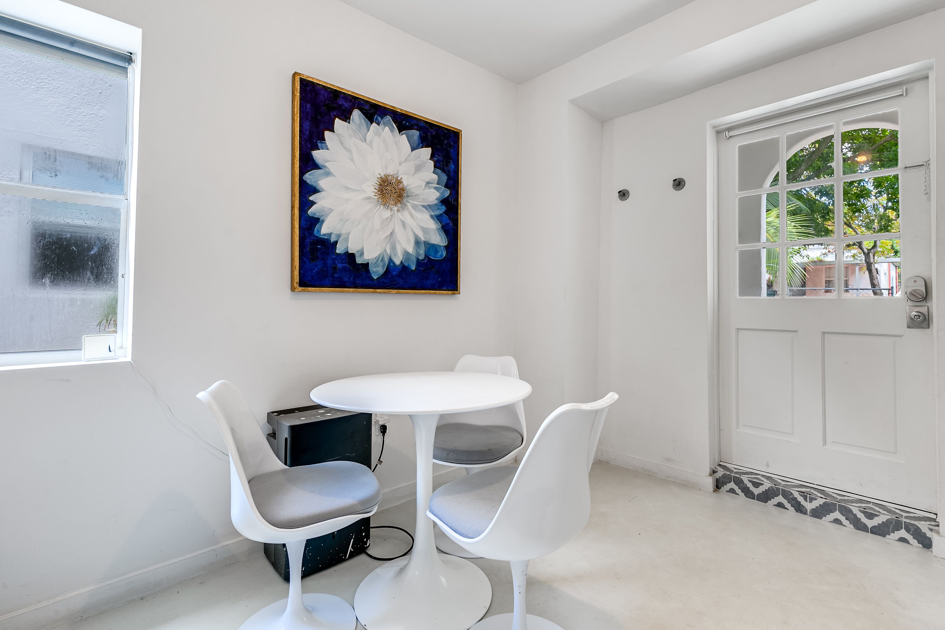 Una habitación con una puerta blanca, una ventana con marco blanco, una mesa blanca con dos sillas y un cuadro grande de flores en la pared.