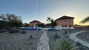 A courtyard with a stone pathway, two buildings, and various plants. - Sunset Village (Luperon)
