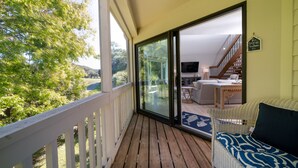 Eine Holzterrasse mit weißer Reling, ein Korbssessel mit gemustertem Kissen und Blick auf ein Wohnzimmer mit Sofa, Couchtisch und Fernseher.