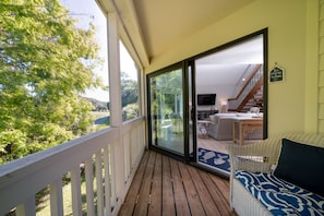 Une terrasse en bois avec une balustrade blanche, un canapé en osier agrémenté d’un coussin à motifs, et une vue sur un salon comprenant un canapé, une table basse et un téléviseur.