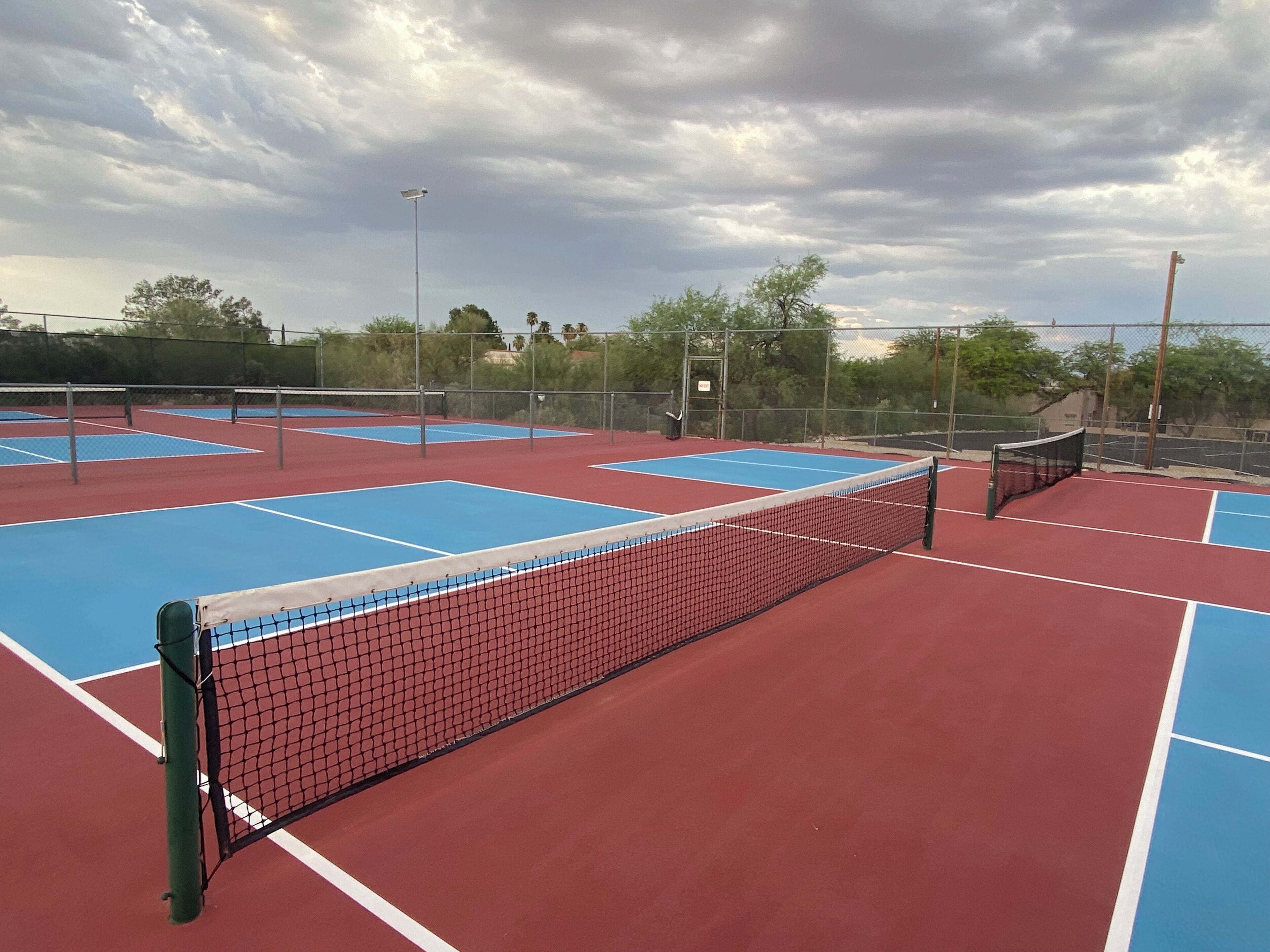 A tennis court with multiple blue and red courts, surrounded by a fence, and a cloudy sky above.