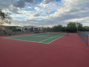 Ein Tennisplatz im Freien mit roter Belag, umgeben von einem Zaun und unter einem bewölkten Himmel.