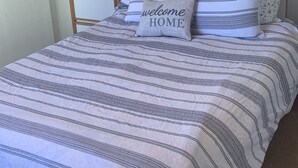 A neatly made bed with striped bedding and a 'welcome home' pillow, a wooden nightstand with a lamp, and a wall-mounted shelf with framed pictures.