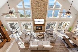 A spacious living room with a stone fireplace, large windows, and a ceiling fan.