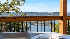 Un bain à remous doté d’une terrasse en bois et de rampes en métal, offrant une vue sur un lac et des collines.