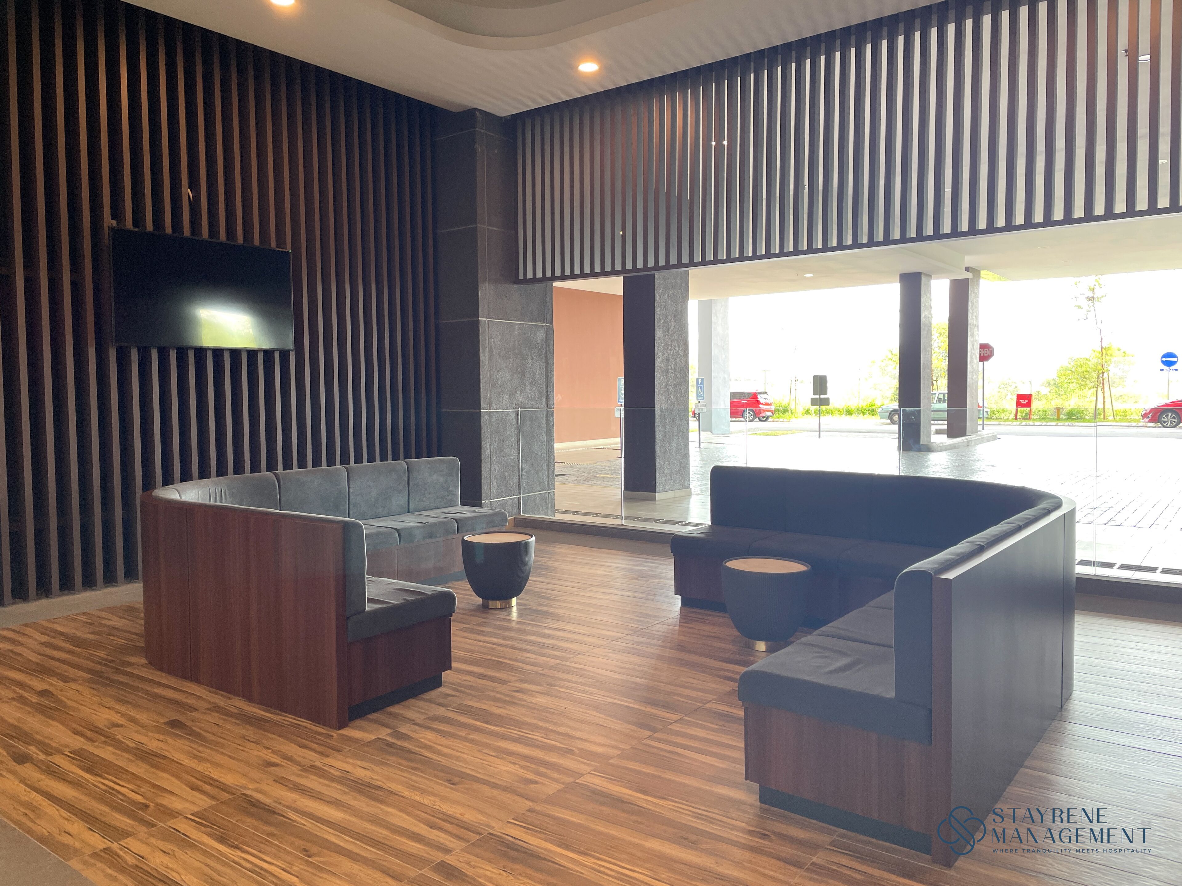 A modern lobby with dark wood flooring, a large sectional sofa, a round black coffee table, and a flat-screen TV mounted on the wall.