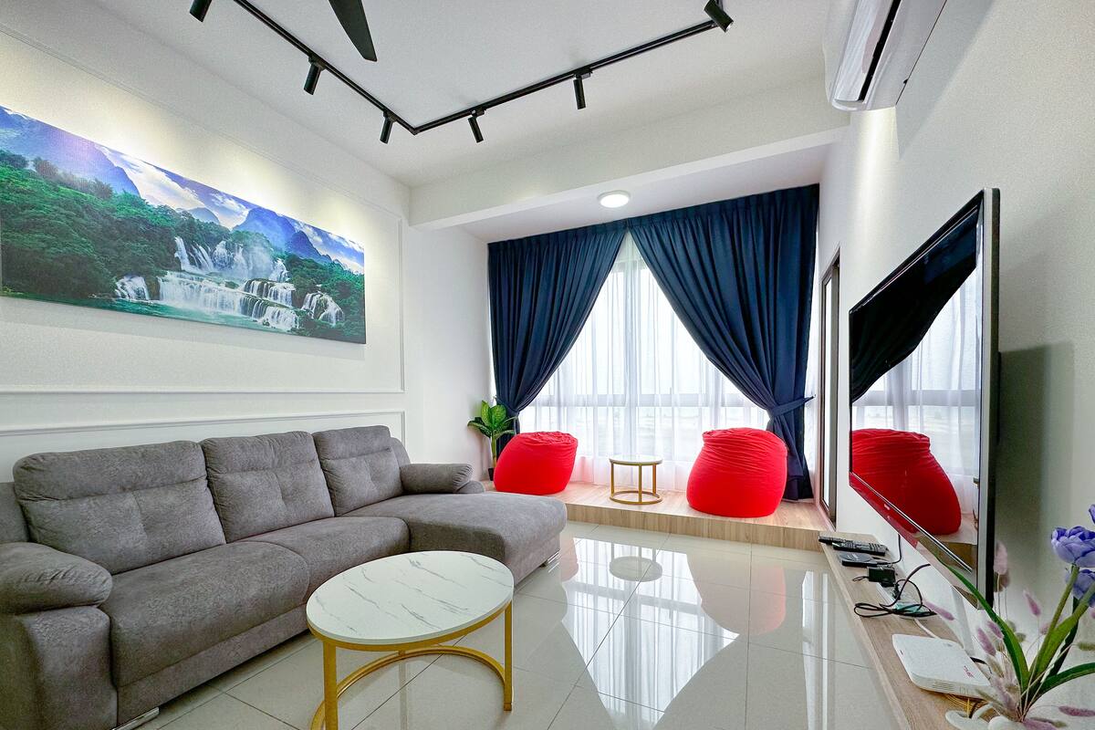 Een moderne woonkamer met een grijze bank, een witte salontafel, rode schapenvachtjes, een grote flatscreen televisie en een schilderij van een waterval aan de muur.