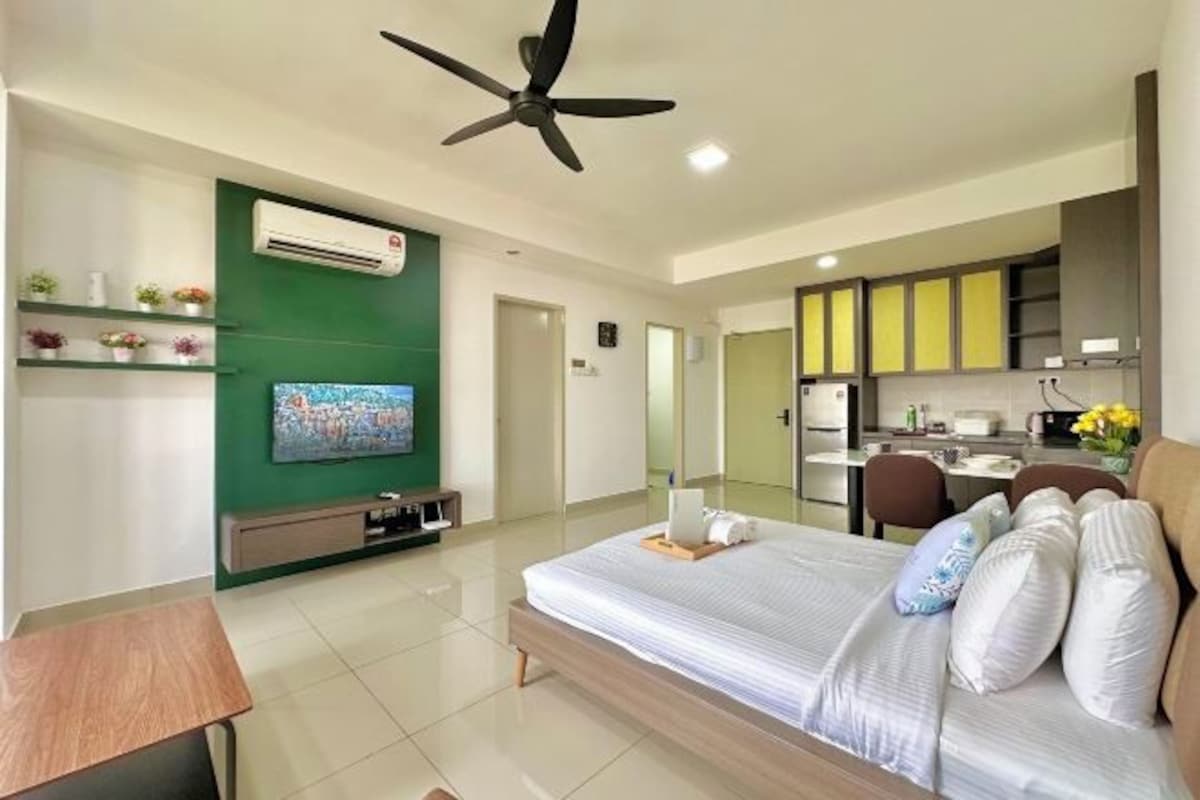 Een moderne hotelkamer met een groot bed, een aan de muur bevestigde televisie, een plafondventilator en een kitchenette op de achtergrond.