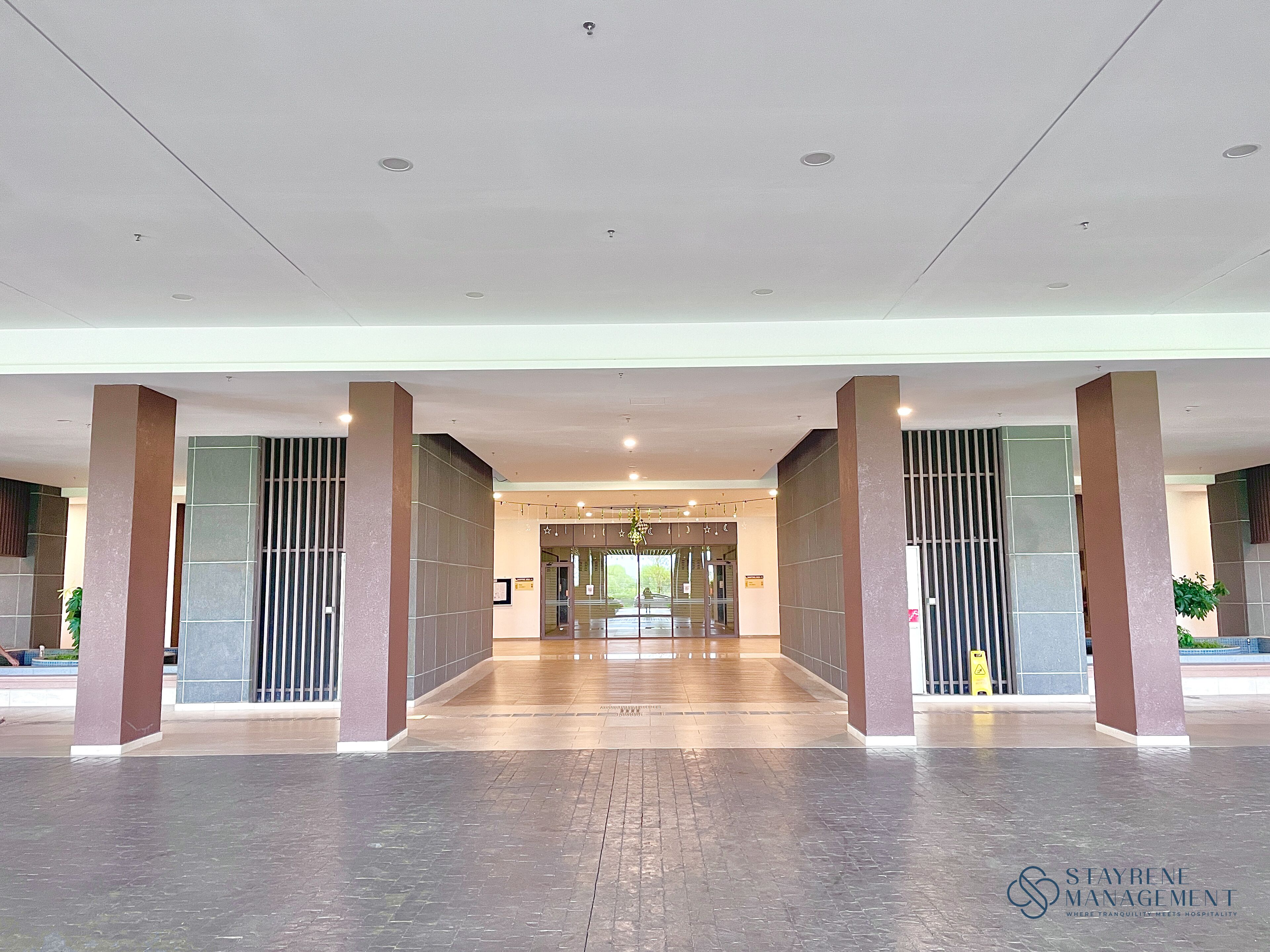 A spacious lobby with a modern design, featuring a central entrance with glass doors, flanked by large pillars, and a tiled floor.