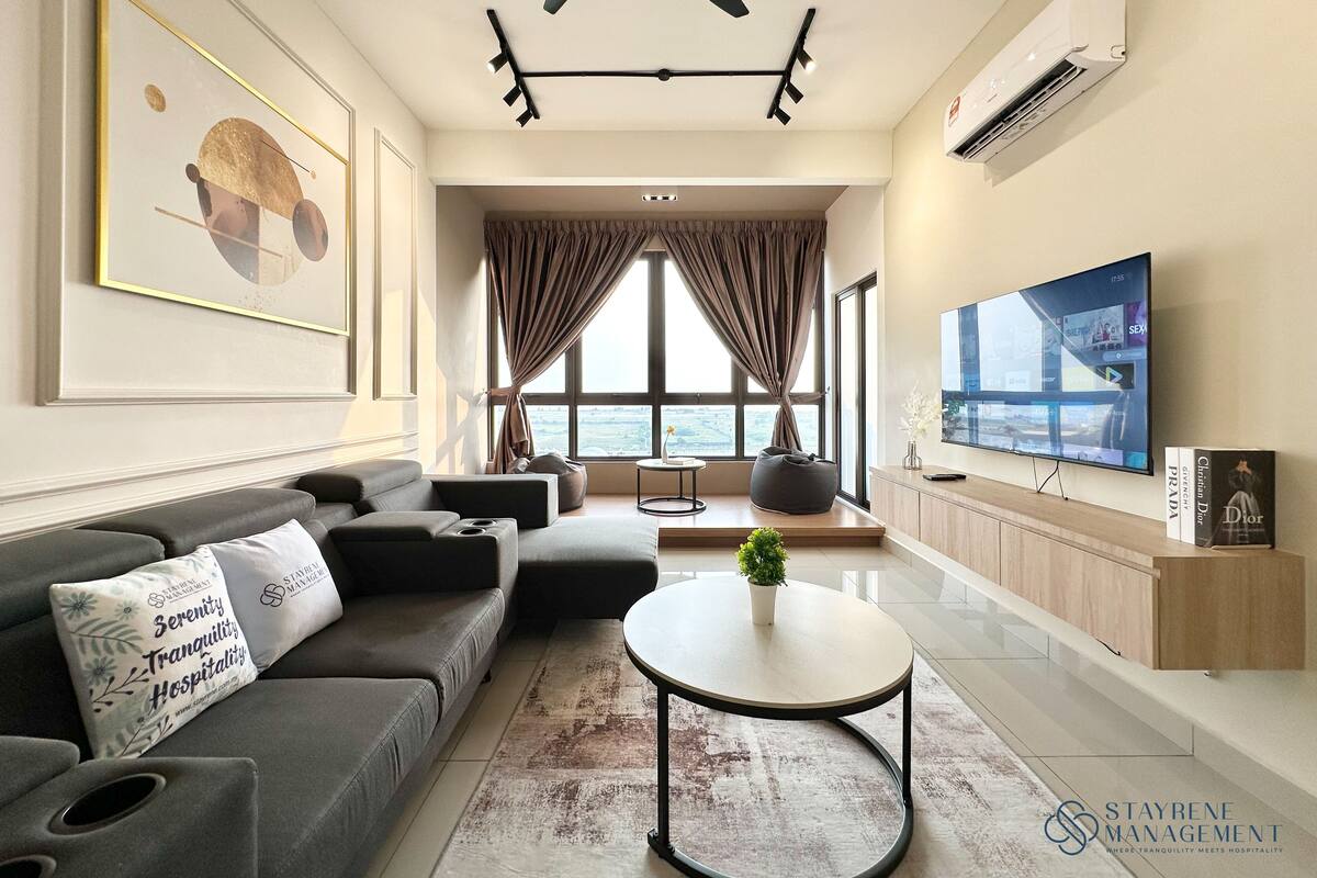 Een moderne woonkamer met een grijze bank, een ronde salontafel, een flatscreen televisie en uitzicht op de stad door grote ramen.