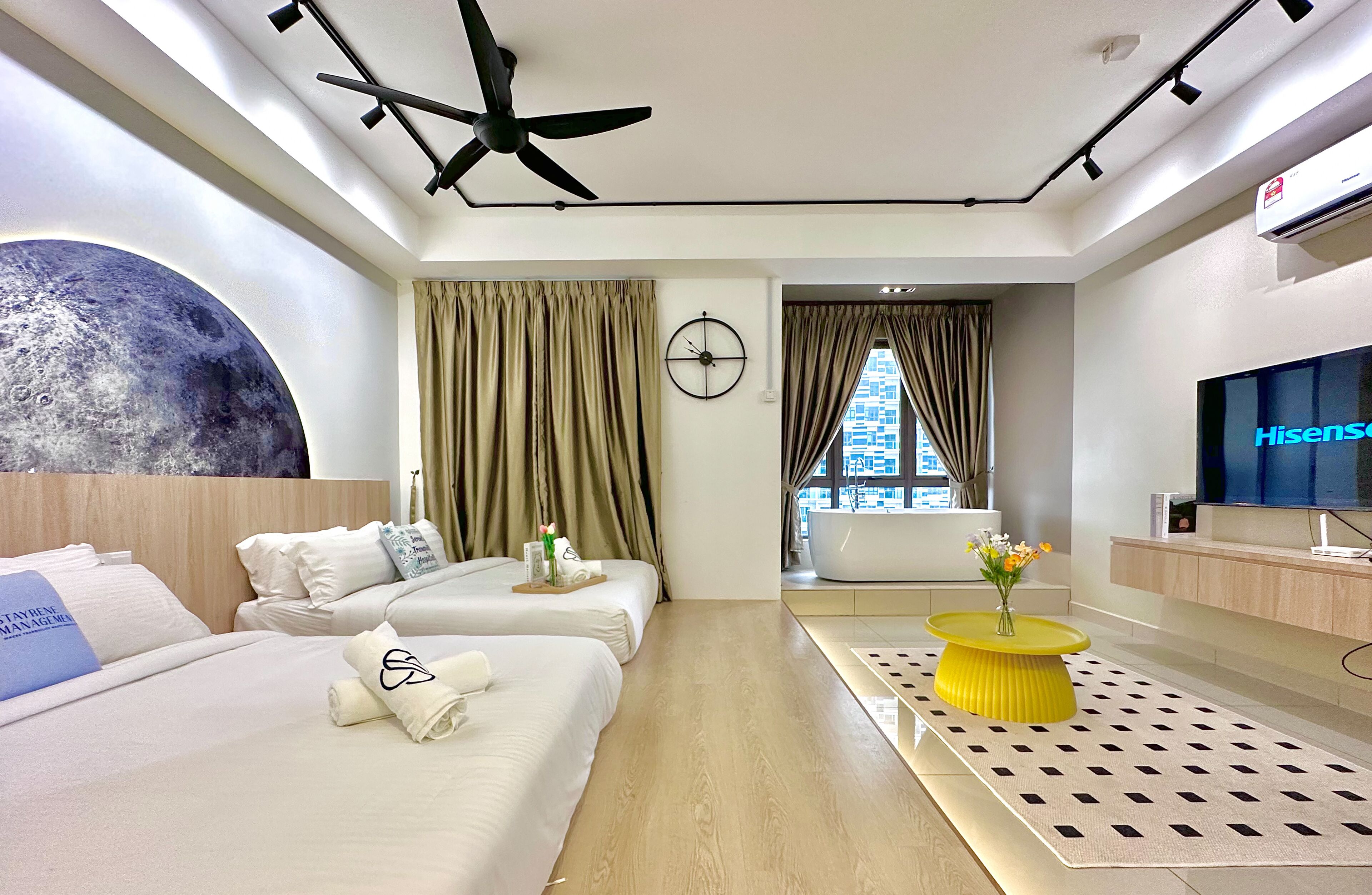 A hotel room with two beds, a ceiling fan, a large wall mural, a TV, and a view of a building through the window.