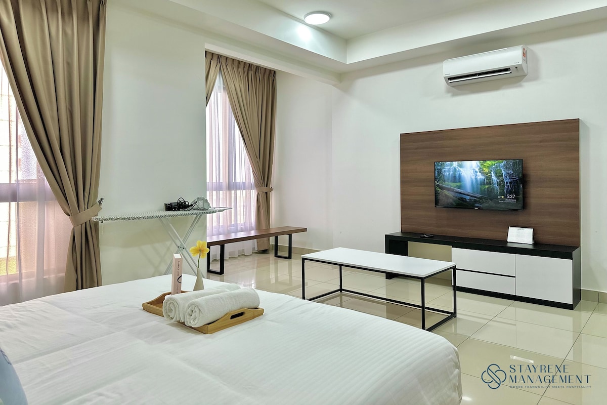 Habitación de hotel con cama, televisor montado en un panel de madera, aire acondicionado, una mesita y una ventana con cortinas.