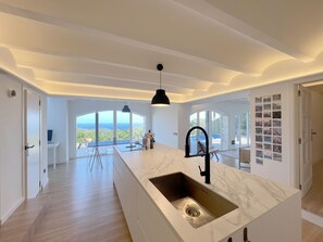 Une cuisine moderne dotée d’un grand îlot central, d’un luminaire suspendu noir et offrant une vue sur l’océan grâce à de grandes fenêtres.