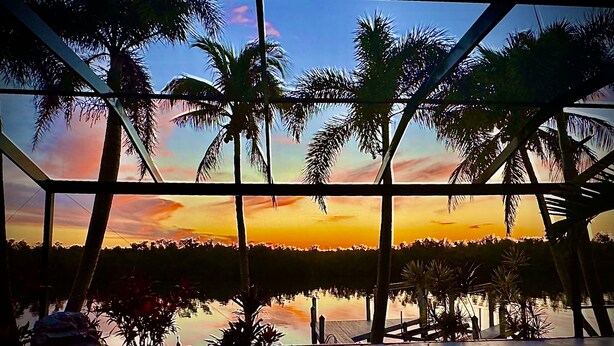 Vista a través de una ventana con atardecer, palmeras y una piscina.
