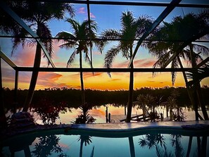 A view through a window with a sunset, palm trees, and a pool. - Completely renovated on 200’ wide spreader canal, backyard sunsets and privacy (Cape Coral)