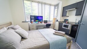 Een slaapkamer met een bed, een bureau met computer, een kleine kitchenette en een raam met zonwering.