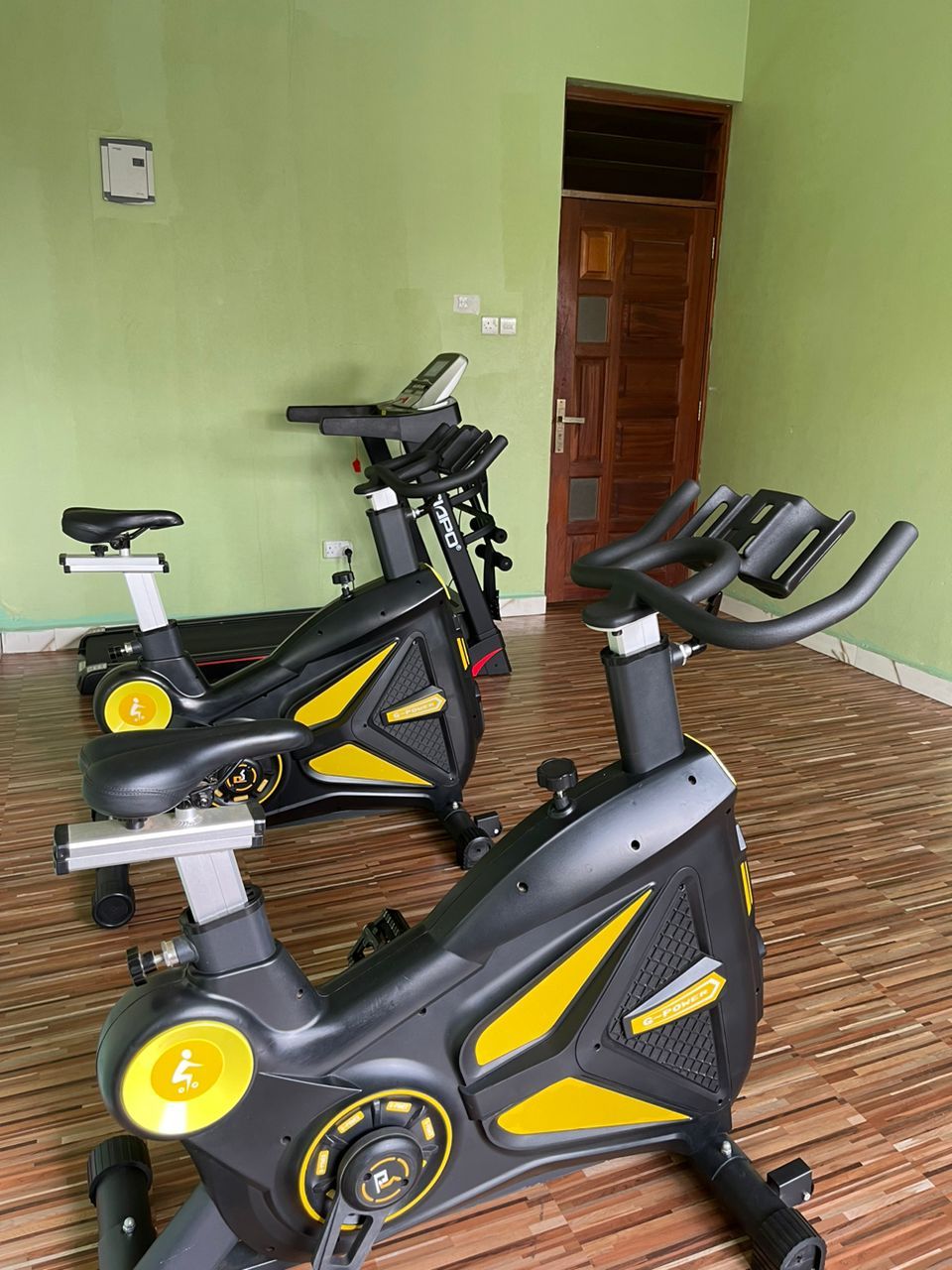 Two stationary bikes in a room with a wooden floor and a green wall.