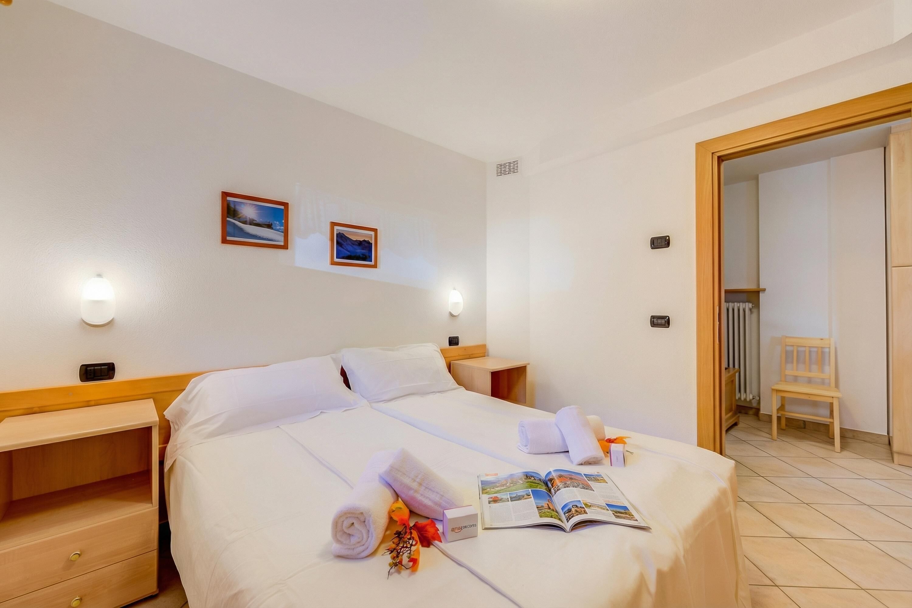 A hotel room with a large bed, two bedside tables, a nightstand, a wooden chair, and a door leading to another room.