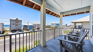 A covered balcony with wooden chairs, a view of the beach, and buildings in the background.
