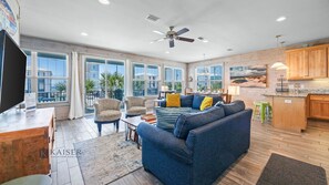 A spacious living room with a large sofa, two armchairs, a dining table, and a kitchen area with wooden cabinets and a countertop.