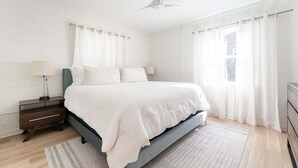 A bedroom with a bed, nightstands, a lamp, a ceiling fan, and a view of the outdoors through a window. - Pet-Friendly St. Simons Island Beach Rental | The Blue Pearl (Saint Simons Island)