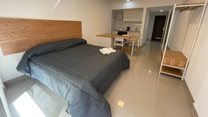 Une chambre à coucher moderne avec un lit, une tête de lit en bois, un bureau et, au fond, un coin cuisine.