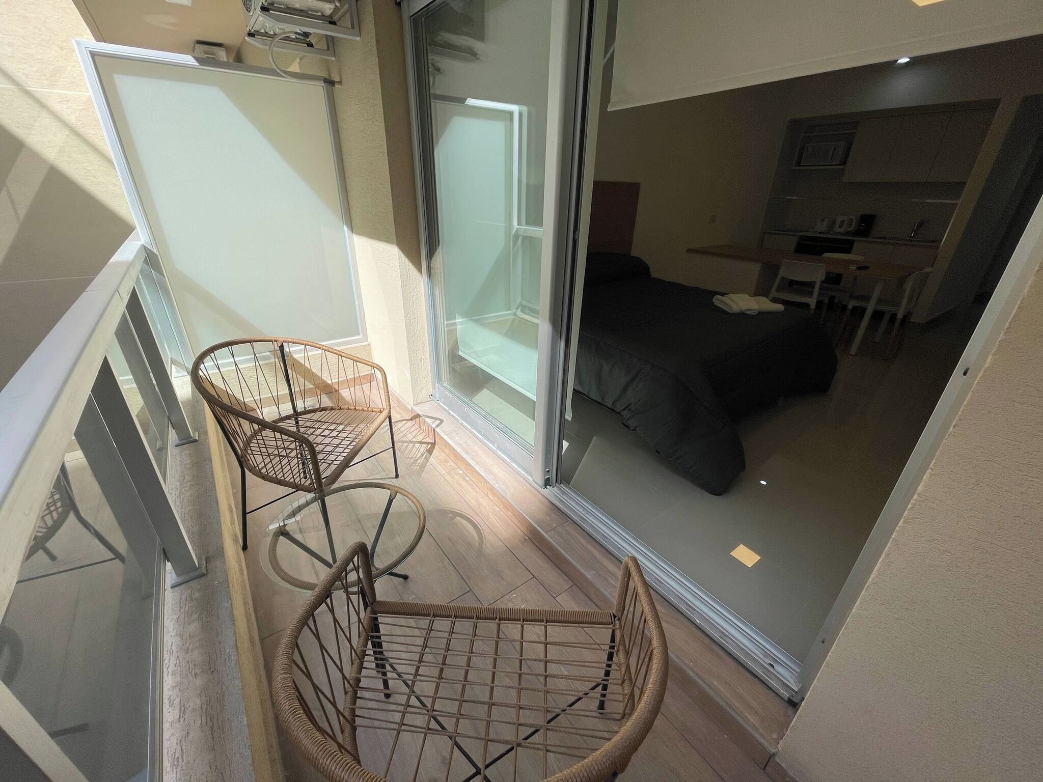 Un balcón con sillas de mimbre, una puerta corrediza de vidrio que da a un dormitorio con cama y una mesita.