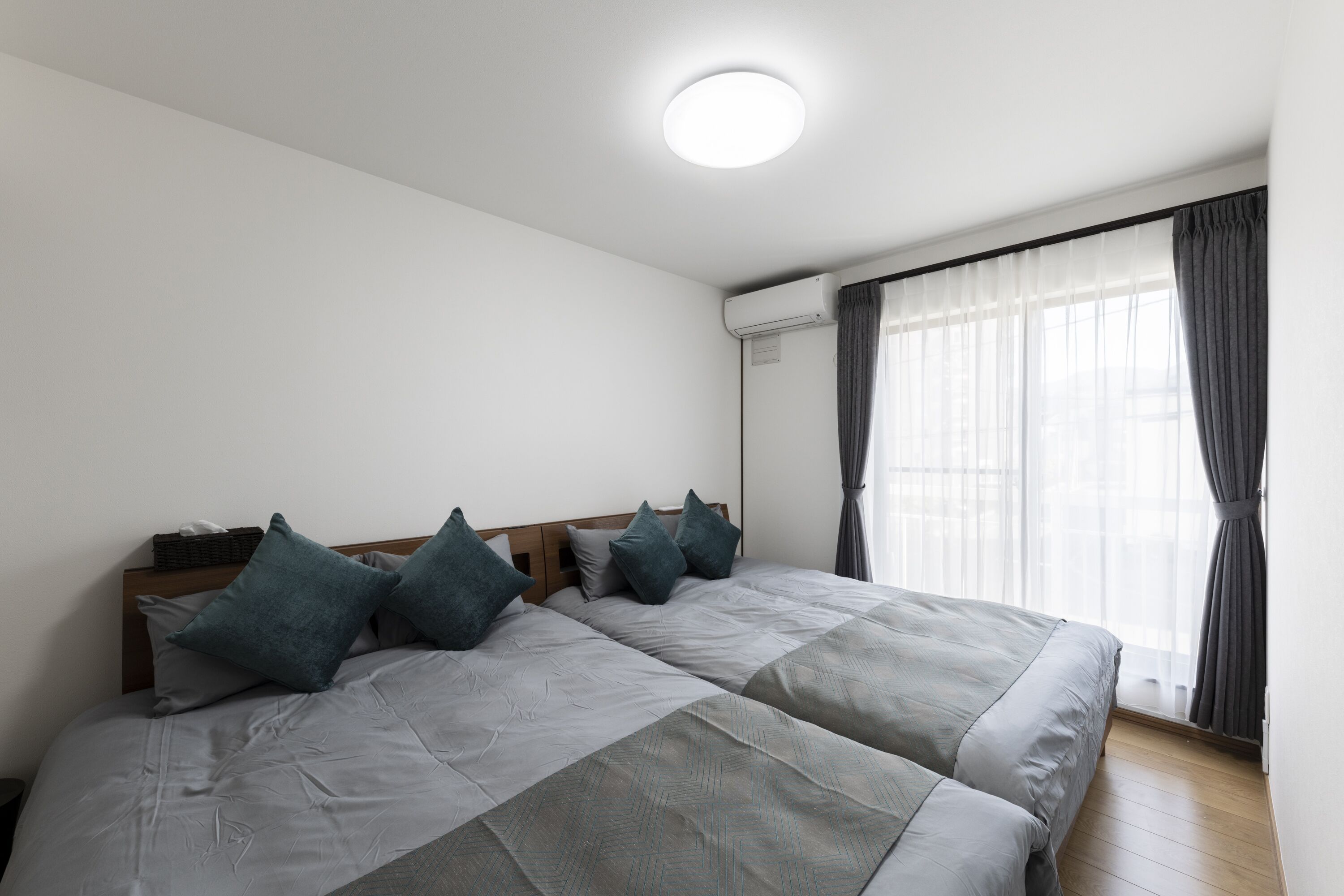 A double bed with grey bedding and teal pillows, a wooden headboard, and a window with sheer curtains.