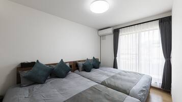 A double bed with grey bedding and teal pillows, a wooden headboard, and a window with sheer curtains.