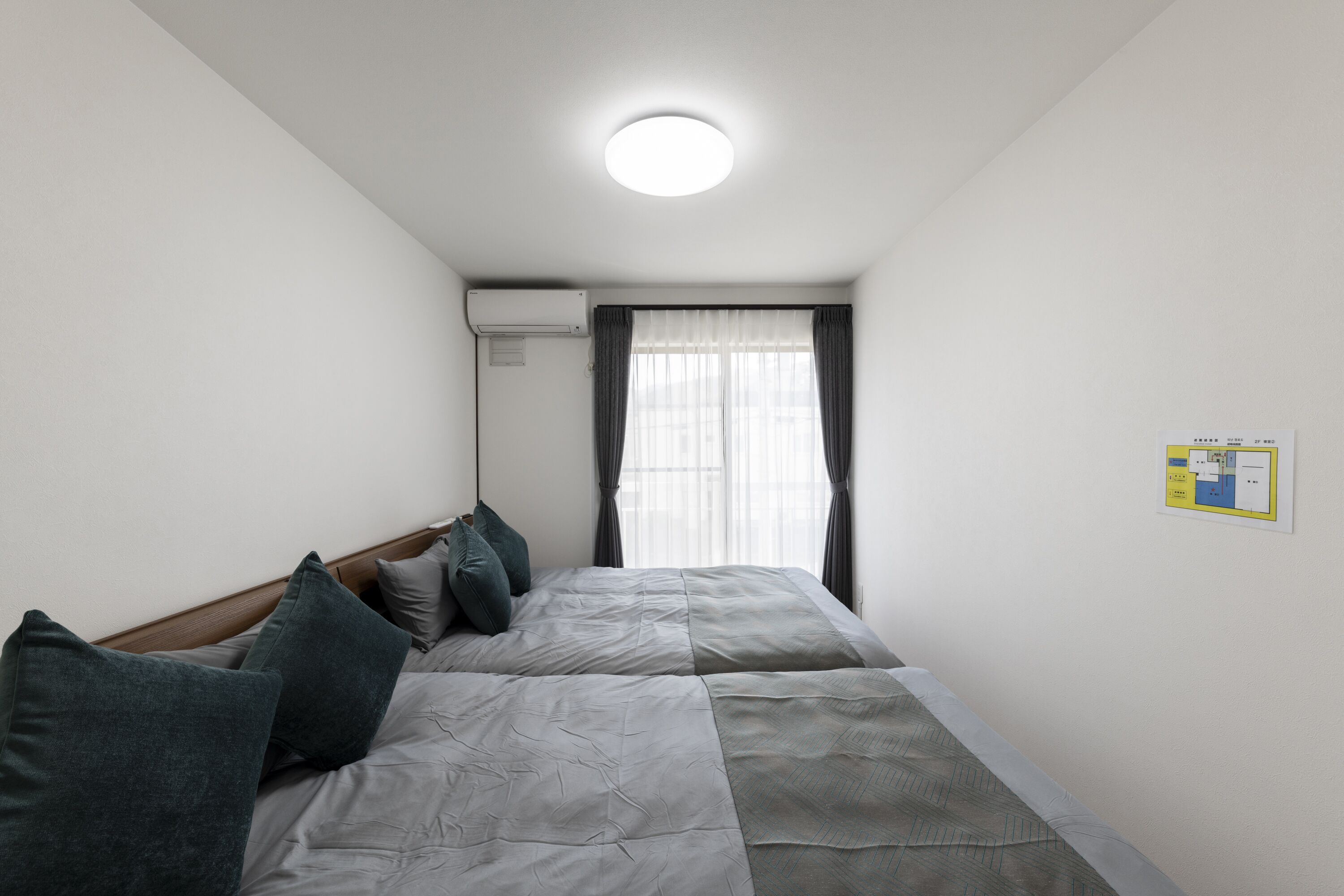 A narrow hotel room with two beds, a ceiling light, an air conditioning unit, and a window with curtains.