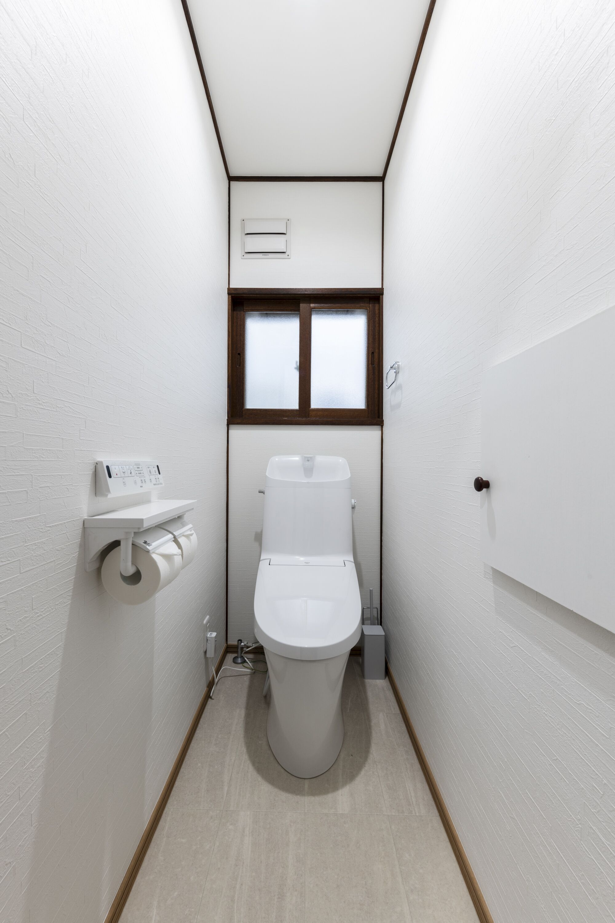 A narrow bathroom with a toilet, a window, and a wall-mounted toilet paper holder.