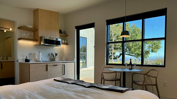 Un dormitorio moderno con una cama grande, un área de comedor con dos sillas, una cocina con armarios de madera y un ventanal con vistas panorámicas.