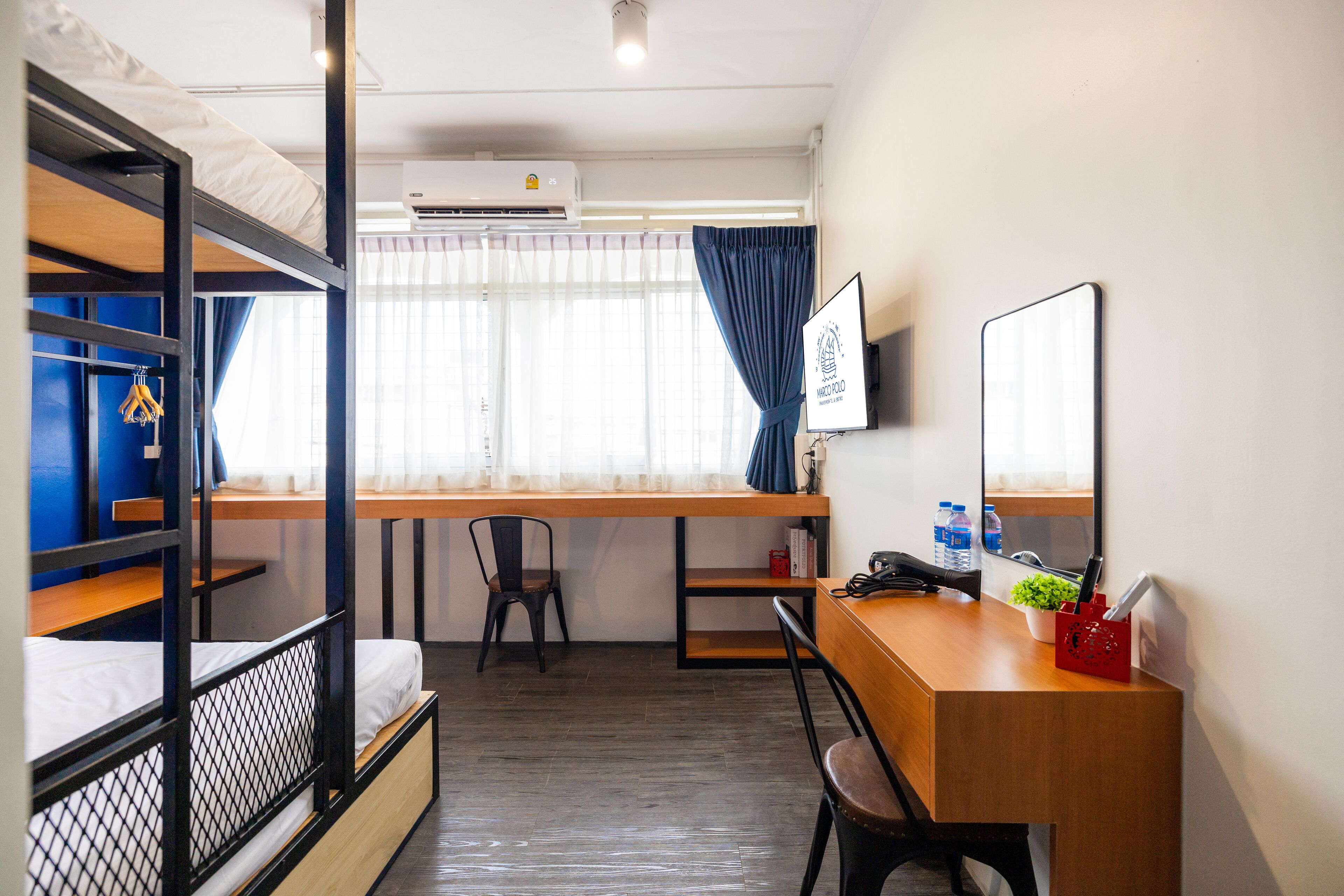 A dorm-style room with bunk beds, a desk, a chair, a TV, and a window with curtains.
