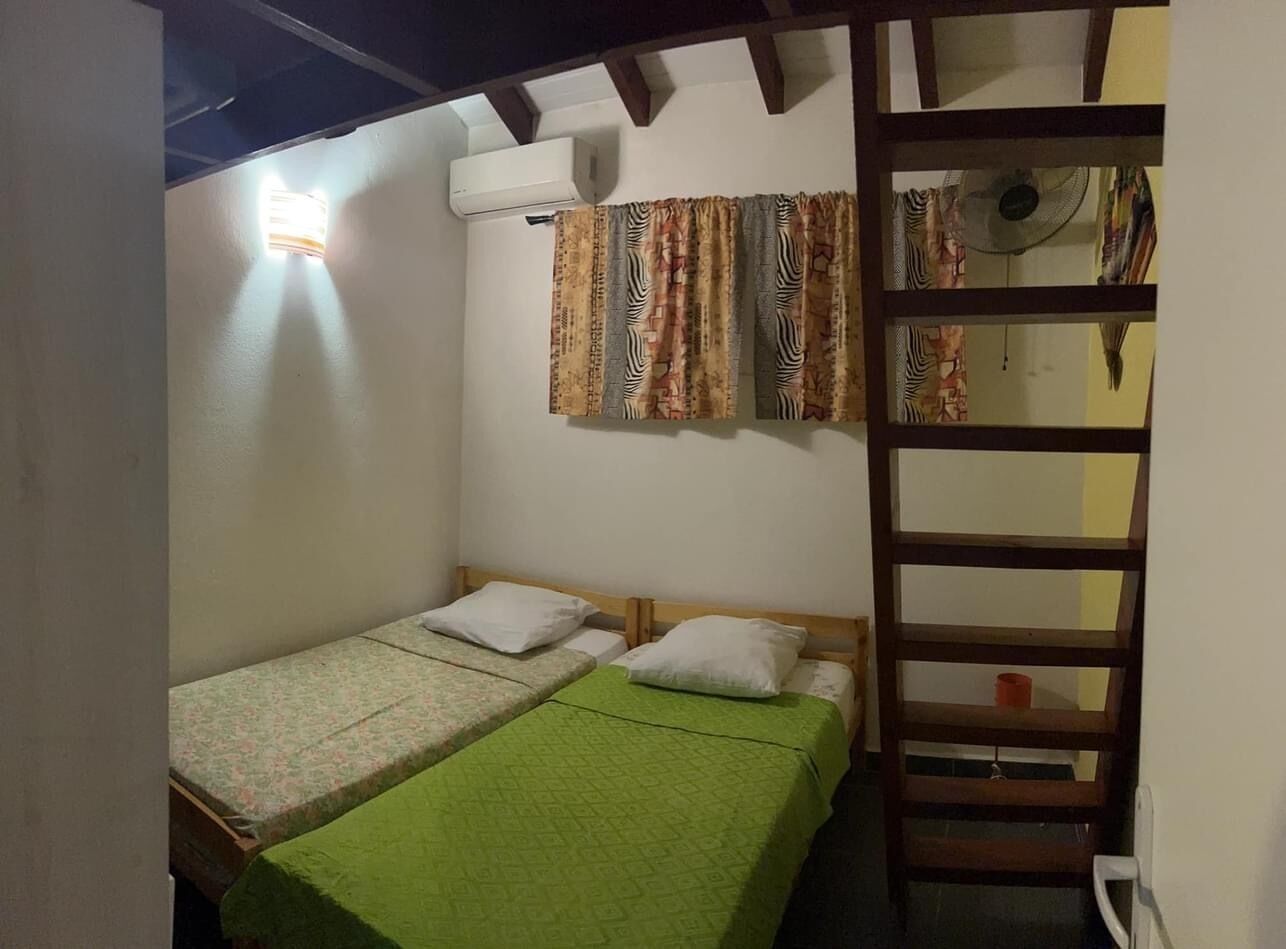 A small, compact room with two single beds, a wooden ladder, and a window with patterned curtains.