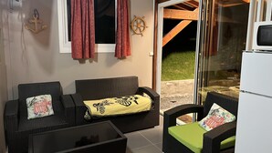 A cozy outdoor seating area with wicker furniture, a glass door leading outside, and a television on a stand. - Havre de Paix - Bungalow de Charme (Baie-Mahault)