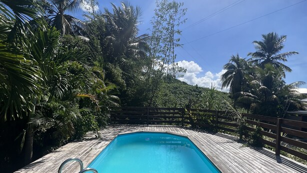 A rectangular swimming pool with a wooden deck surrounding it, set against a backdrop of lush greenery and a clear blue sky. - Havre de Paix - Bungalow de Charme (Baie-Mahault)