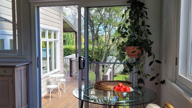 A glass-enclosed patio with a table and chairs, a potted plant, and a view of a garden and house.