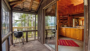 A wooden deck with a grill, a sliding glass door leading to a kitchen, and a view of trees. - Meerman Camp - PRIVATE Rangeley Lake waterfront cottage, with a small beach! (Rangeley)