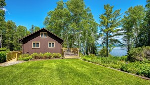 A two-story cabin with a deck, surrounded by a well-maintained lawn and dense trees, overlooking a body of water. - Meerman Camp - PRIVATE Rangeley Lake waterfront cottage, with a small beach! (Rangeley)