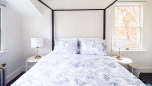 Camera da letto con un letto posizionato al centro sotto una trave a soffitto nera, affiancato da due comodini con lampade, una finestra con vista sugli alberi e un termosifone sulla parete sinistra.