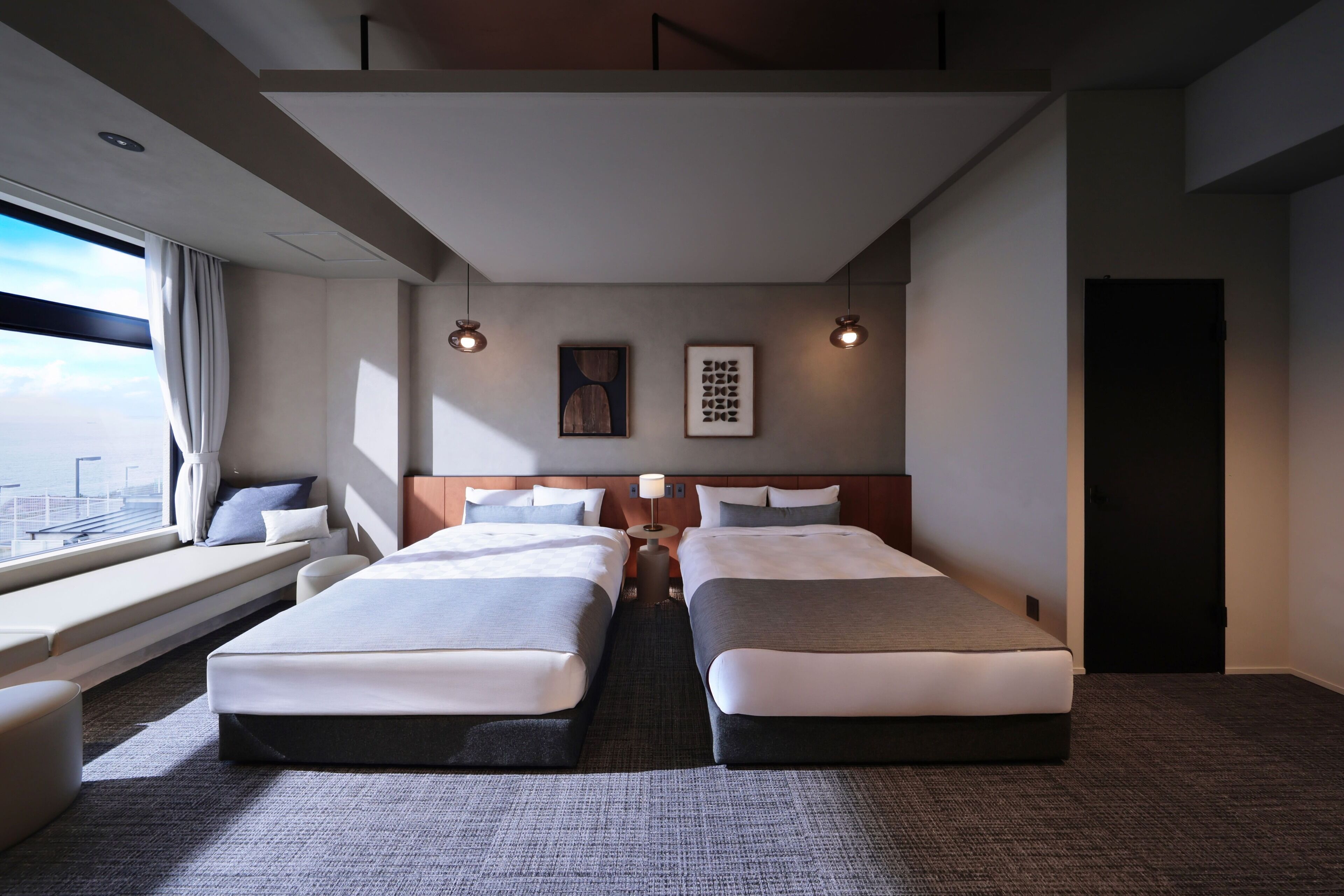Una habitación de hotel con dos camas, un asiento junto a la ventana, vistas a la ciudad y una puerta.