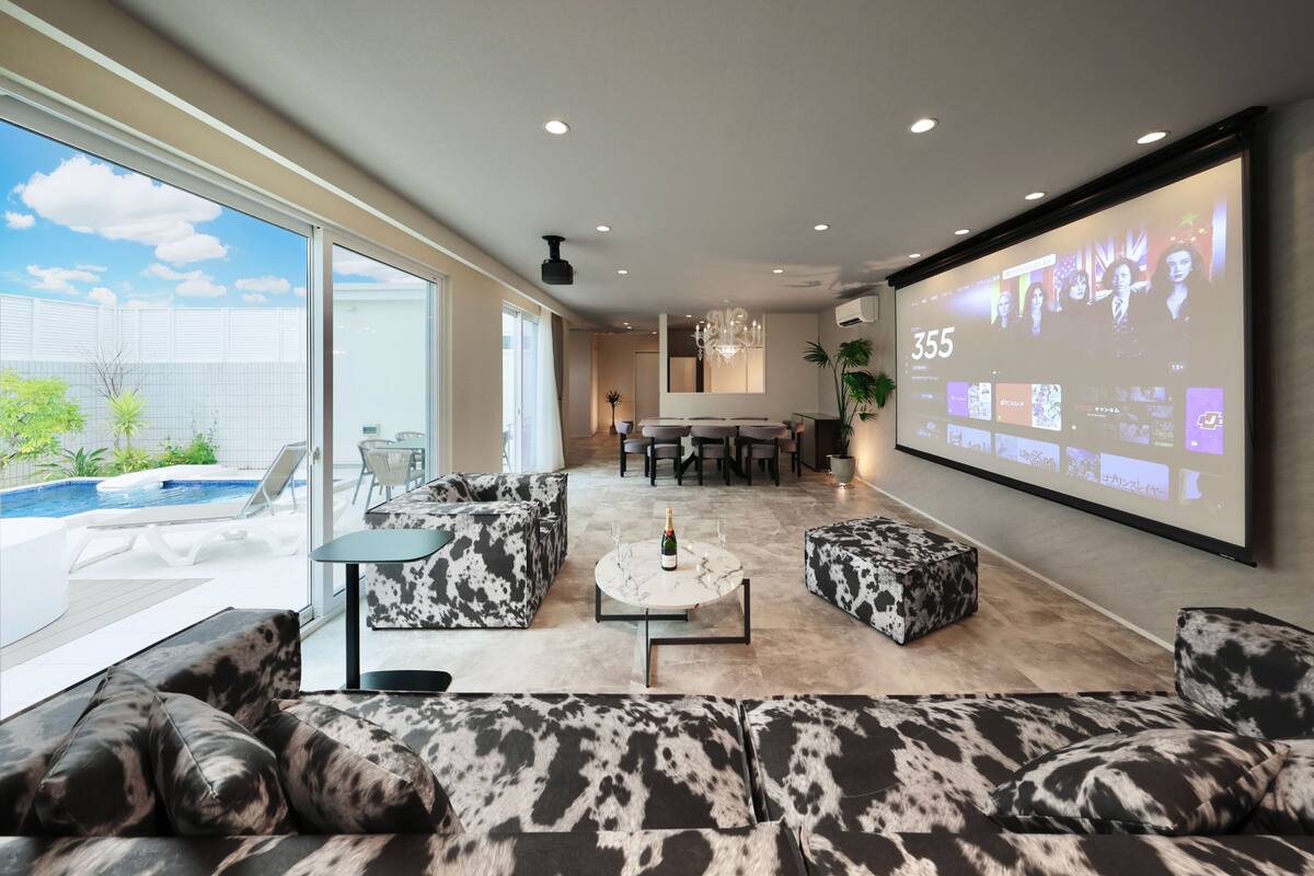 Un salon moderne doté d’un grand écran diffusant une présentation, d’une piscine à l’extérieur et d’un coin salon confortable agrémenté de coussins à motifs.