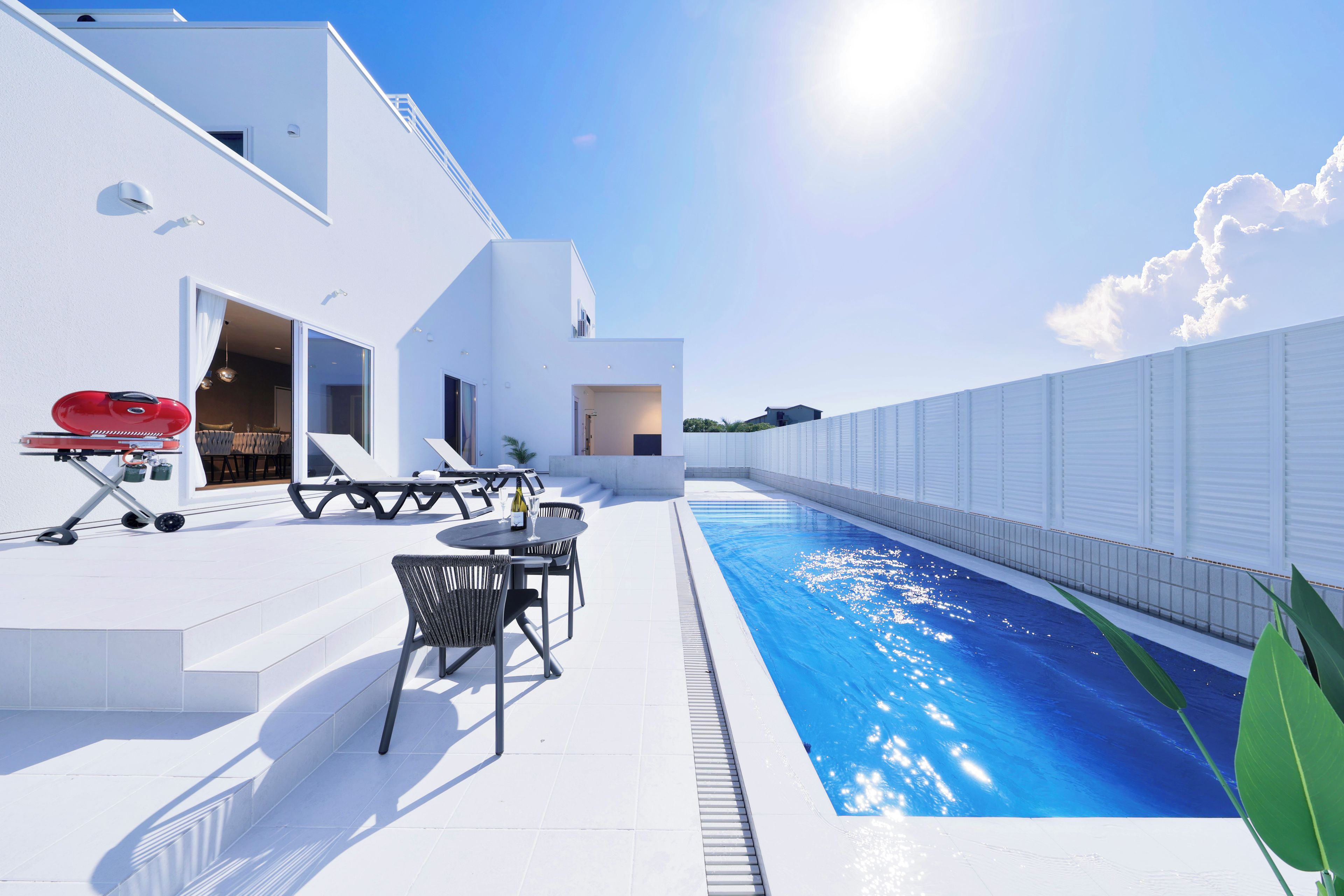 Un área de piscina moderna al aire libre con piscina azul cristalina, zona de barbacoa y espacio de estar con sillas y mesa.