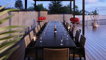 Een buitenterras met een lange tafel gedekt voor een maaltijd, omringd door rieten stoelen, een barbecue en een fles champagne.