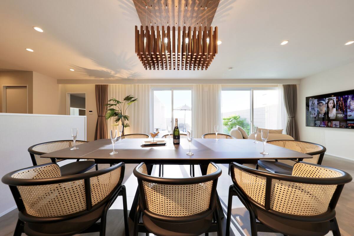 Un espace de salle à manger moderne, avec une longue table, six chaises en osier et un luminaire original en bois suspendu au plafond.