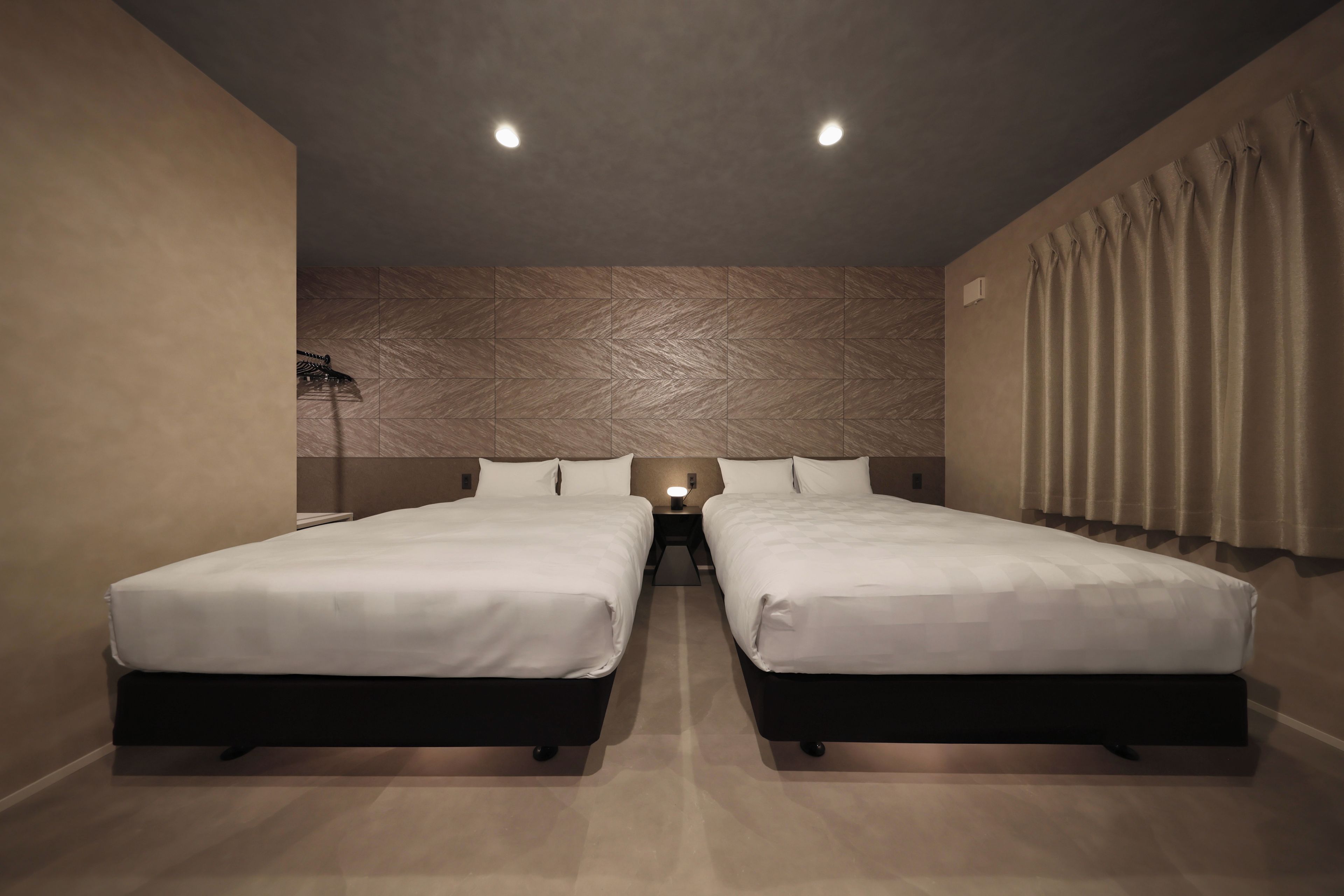 A hotel room with two beds, a textured wall, a nightstand with a lamp, and a curtain.