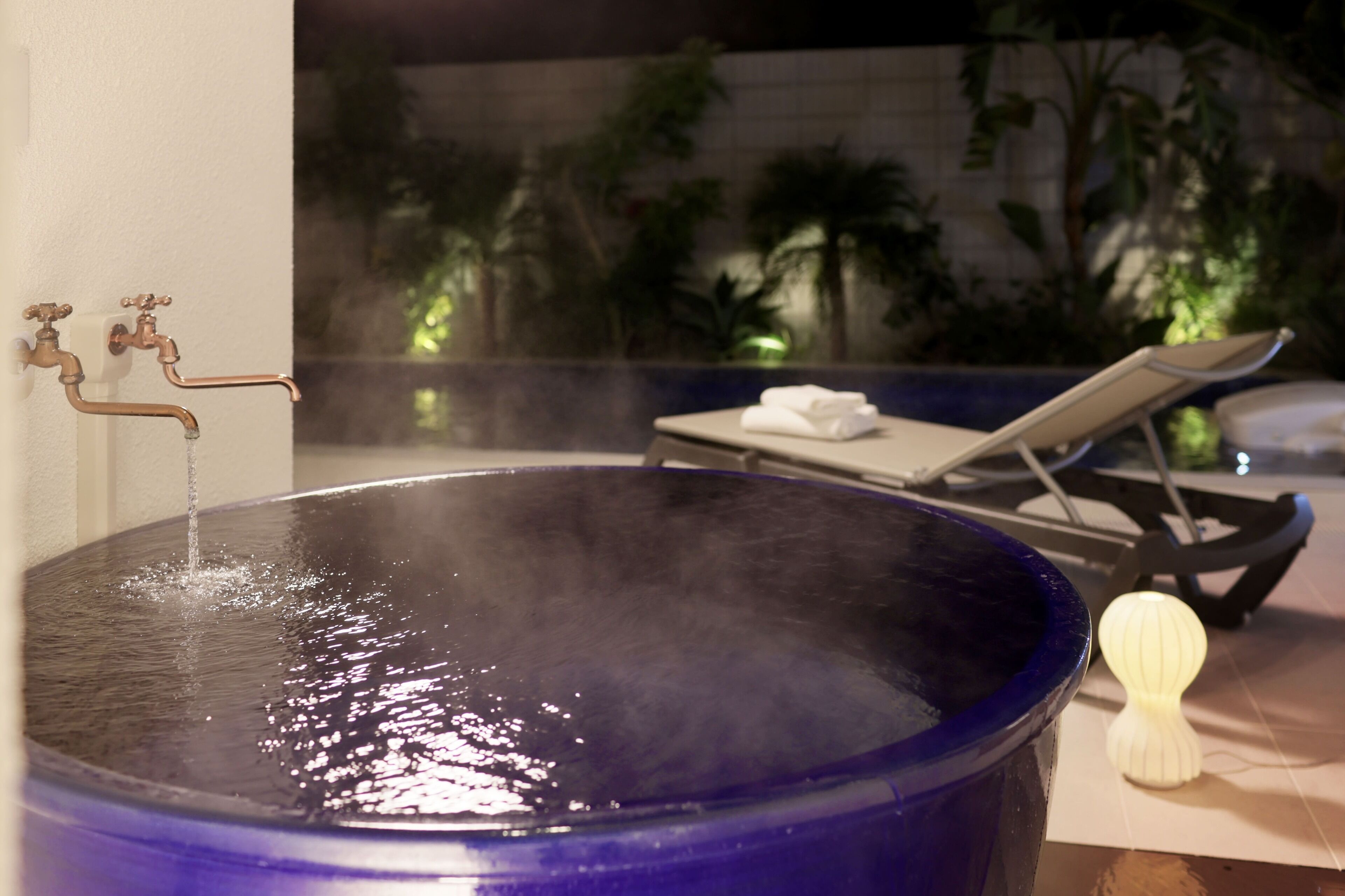 A spa area with a large blue bathtub, a lounge chair, and a small white candle.