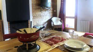 Een eethoek met een houten tafel, een rode pan op een standaard, een aan de muur bevestigde televisie en een raam met gordijnen.
