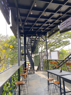 An outdoor seating area with tables and chairs, a staircase, and a view of trees and buildings. - DJURAGAN KAMAR SUHAT (Malang)