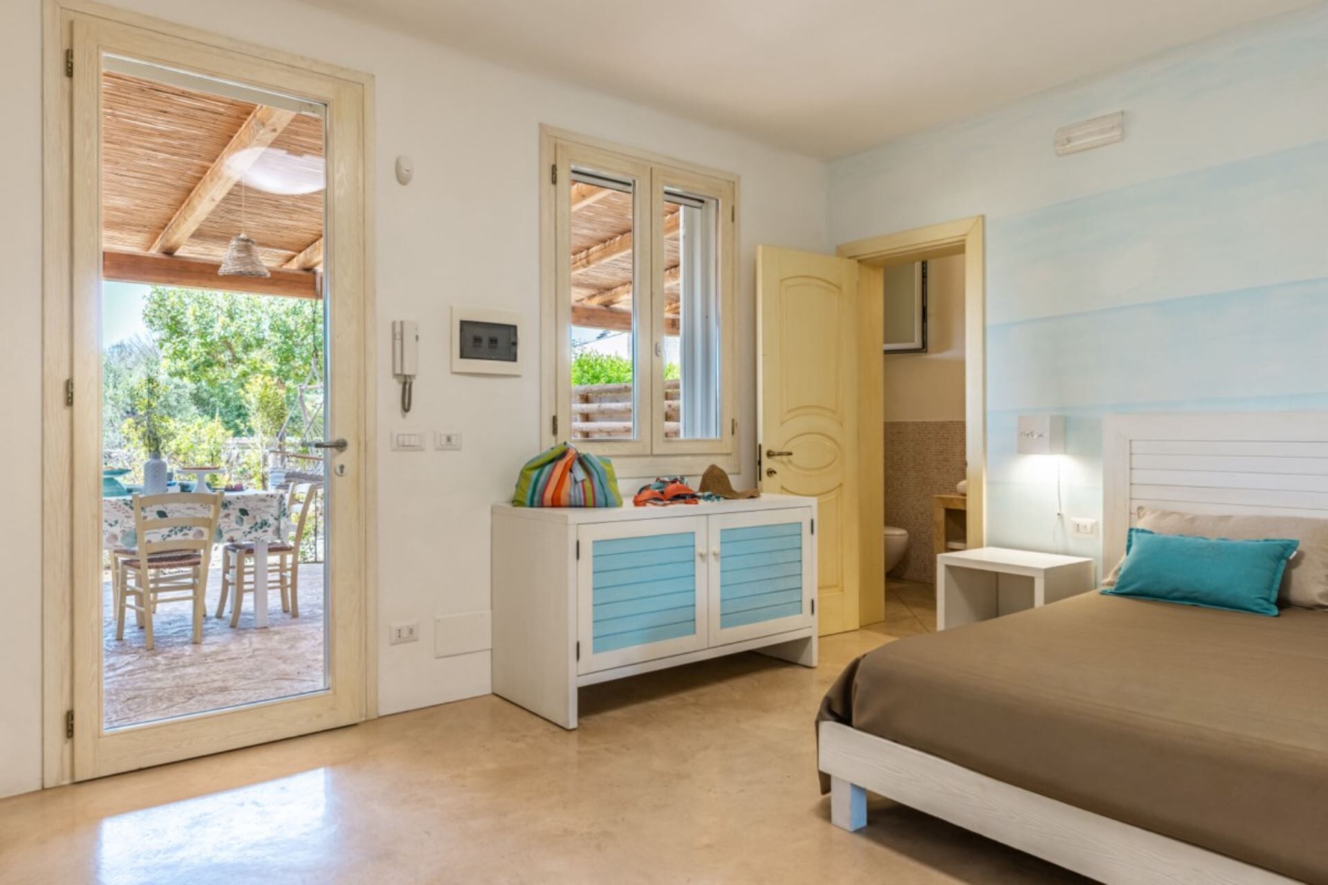 A bedroom with a bed, a bedside table, a wardrobe, a window, and a door leading to an outdoor area with a table and chairs.